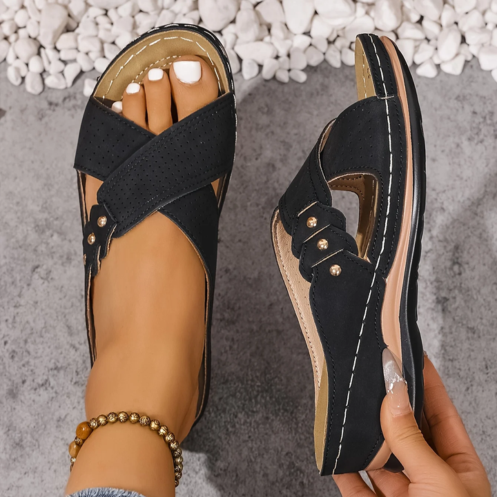 Black orthopedic wedge sandals held in hand, shown on a textured surface with white stones.
