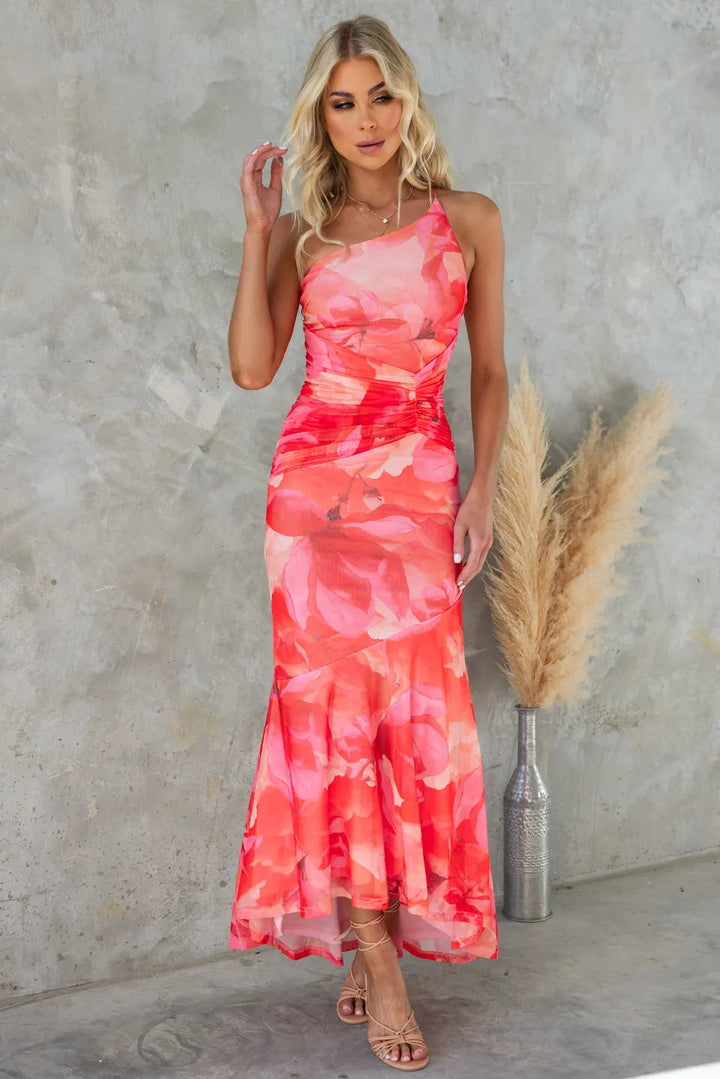One-shoulder coral pink pleated floral dress in a textured studio setting with a gray wall and pampas grass vase.