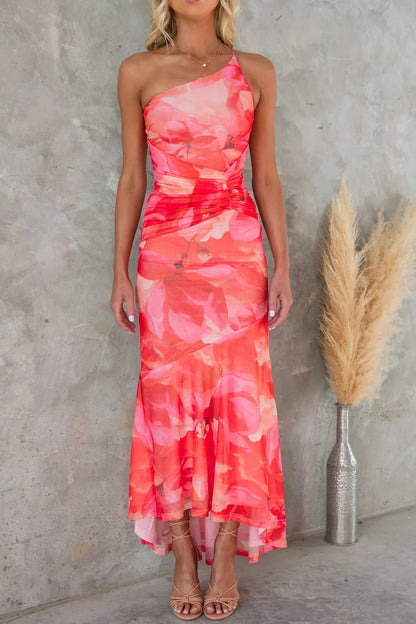 Coral pink floral pleated dress with one shoulder design, set against a gray studio backdrop with pampas grass.