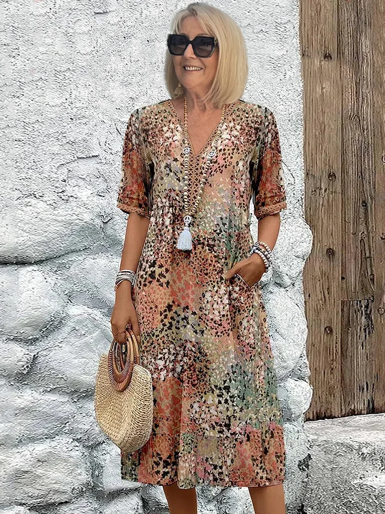 Woman in an earthy-toned midi dress with floral designs, holding a straw handbag, posed against a rustic textured background.