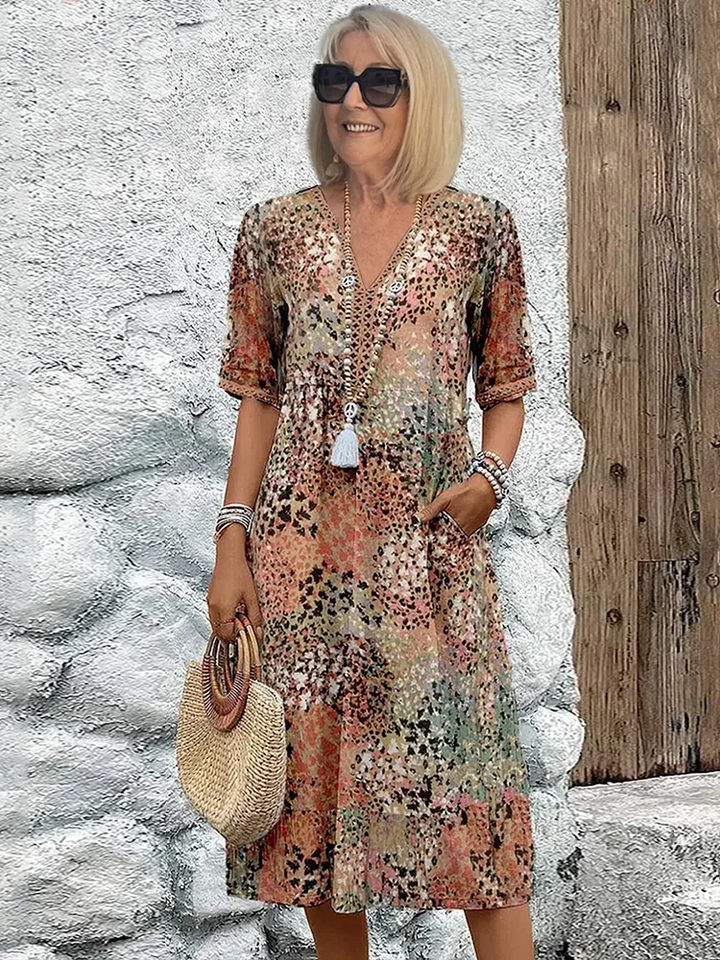 Woman in an earthy-toned midi dress with floral designs, holding a straw handbag, posed against a rustic textured background.