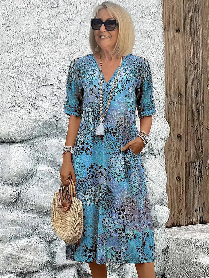 Woman in a blue floral midi dress with accessories, holding a straw handbag, posed against a textured rustic backdrop.