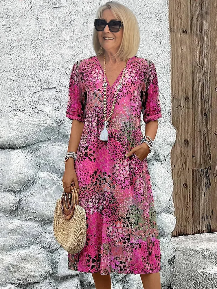 Woman wearing a vibrant pink midi dress with a floral pattern, accessorized with a straw handbag and jewelry, standing against a textured wall.