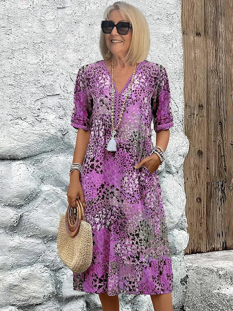 Lady wearing a purple floral midi dress with accessories, holding a straw handbag, standing against a textured rustic background.