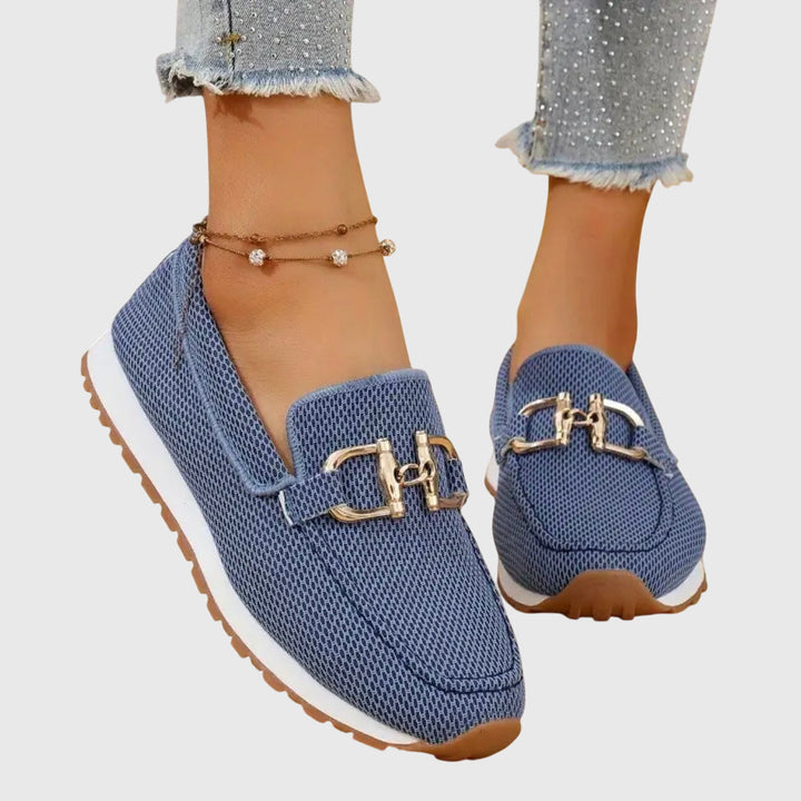 Blue orthopedic loafers with gold accents worn with frayed jeans and anklet on a light background.