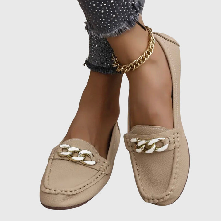 Beige orthopedic loafers with gold and white chain detail, worn with dark pants on a gray background.