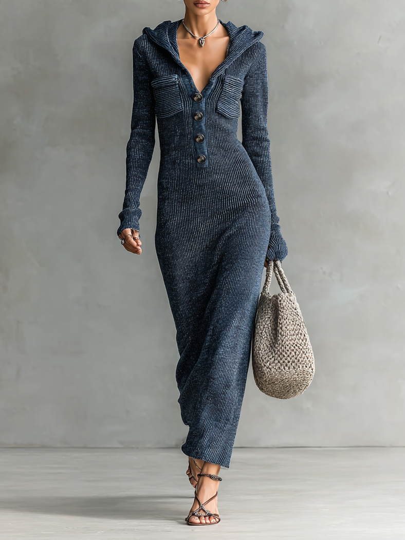 Woman wearing a dark blue slim-fit hooded maxi dress with button detail, standing against a neutral background holding a woven bag.