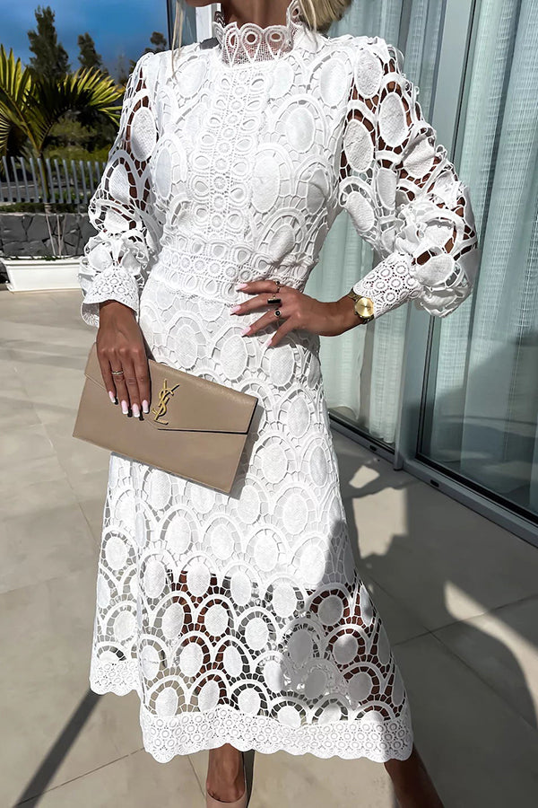 White lace dress featuring detailed patterns and long sleeves, accessorized with a beige clutch, studio backdrop with a glass window.
