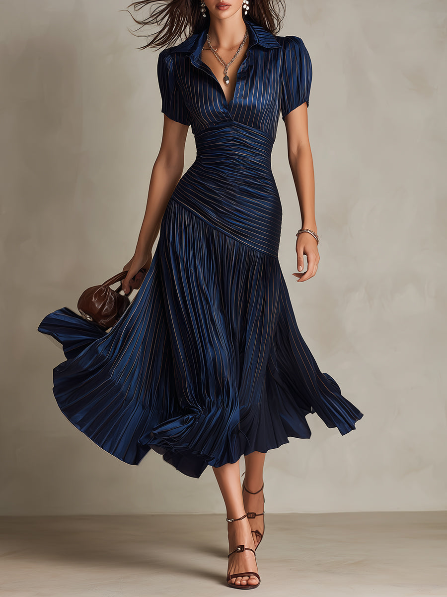 Model in flowing navy midi dress with pleated skirt and short sleeves, holding a purse in a studio setting.