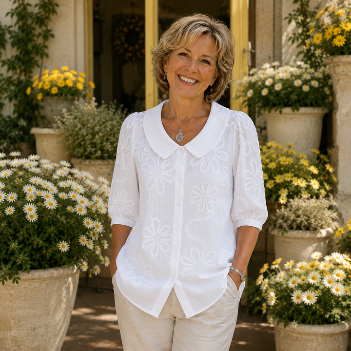 Woman in a white floral blouse poses in garden setting with yellow and white flowers.