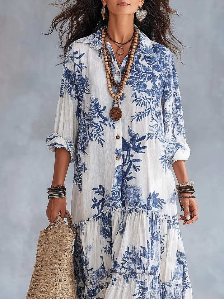 White maxi dress with blue floral print, accessorized with layered necklaces and bracelets, holding a woven bag in studio light.