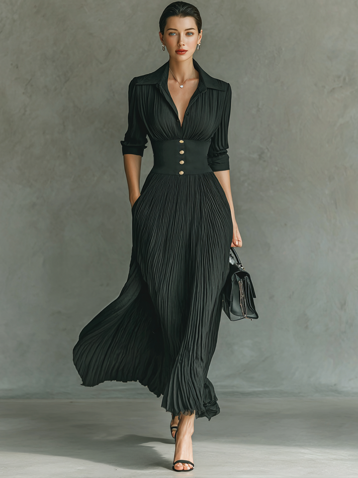 Model wearing a black pleated maxi dress with buttons, standing against a textured gray background, holding a handbag.