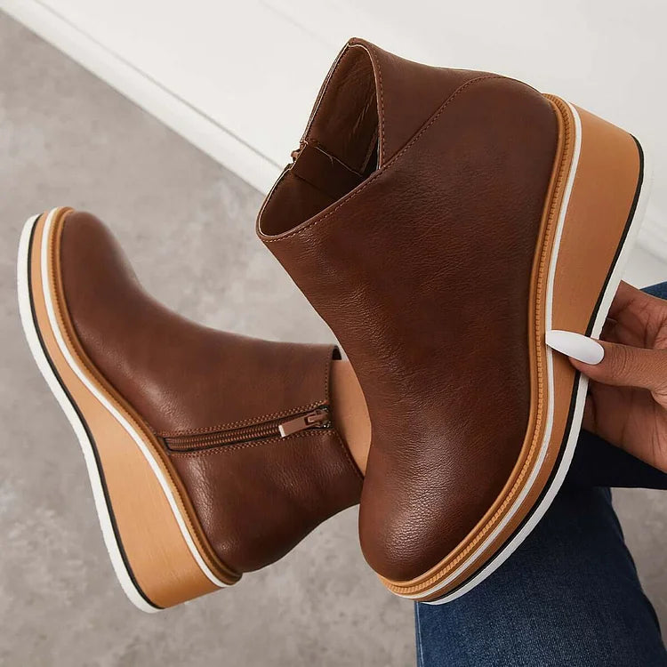 Hand holding a brown boot with a wedge sole and zipper against a gray background.