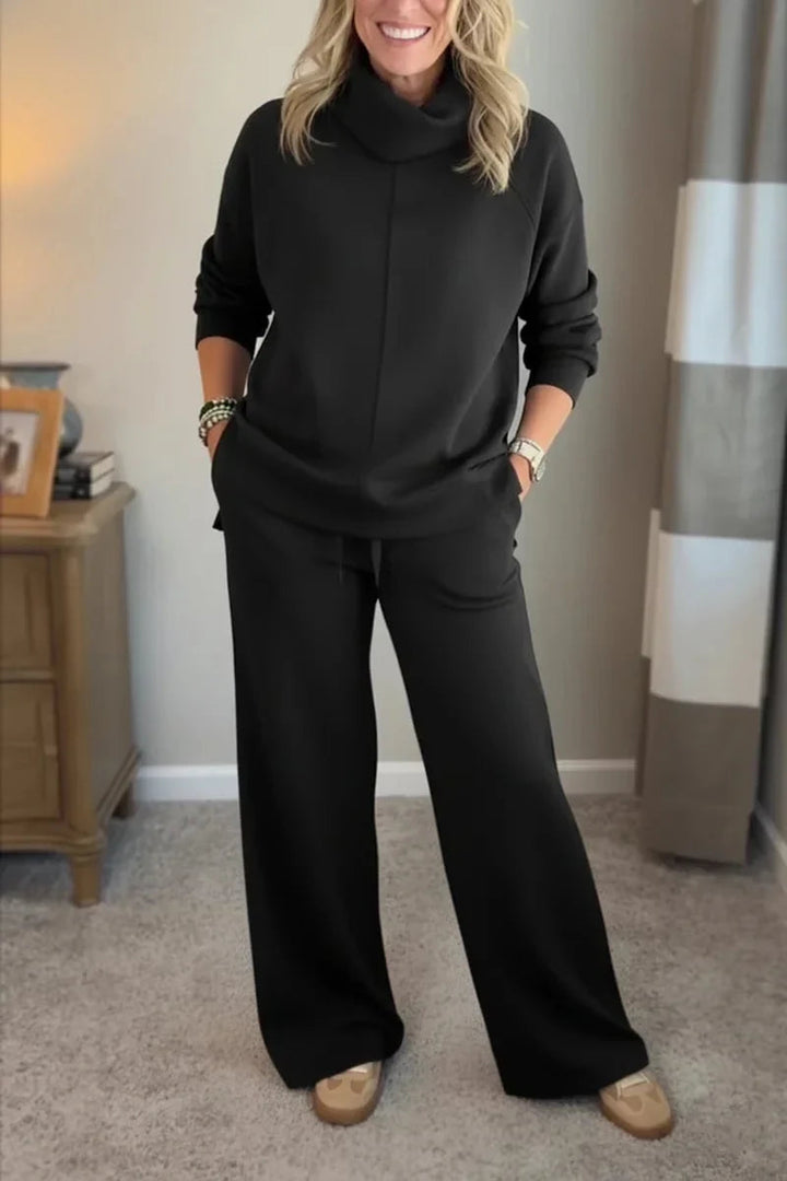 Woman in a black comfy set with long sleeves and wide-leg pants, standing in a carpeted room with a wooden table nearby.
