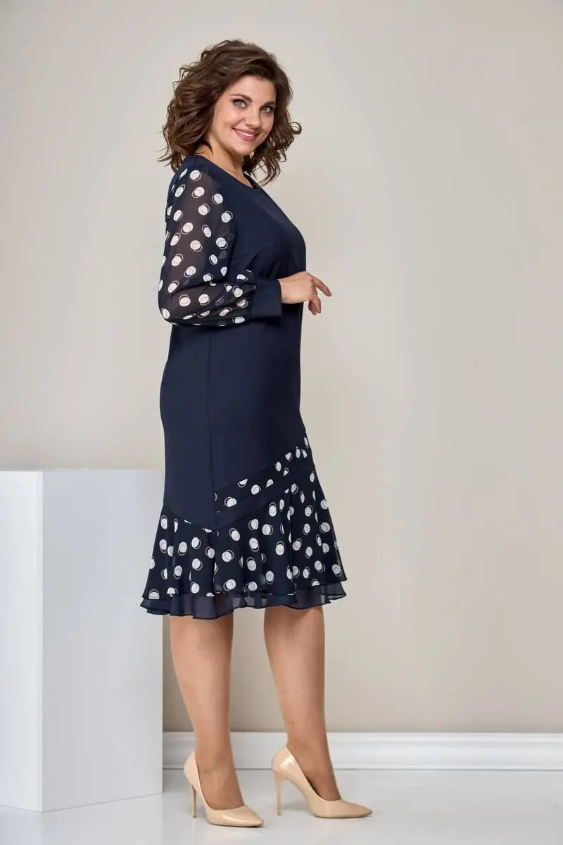 Side view of woman in navy dress with polka dot sleeves, asymmetric ruffled hem, beige heels, in a neutral room.