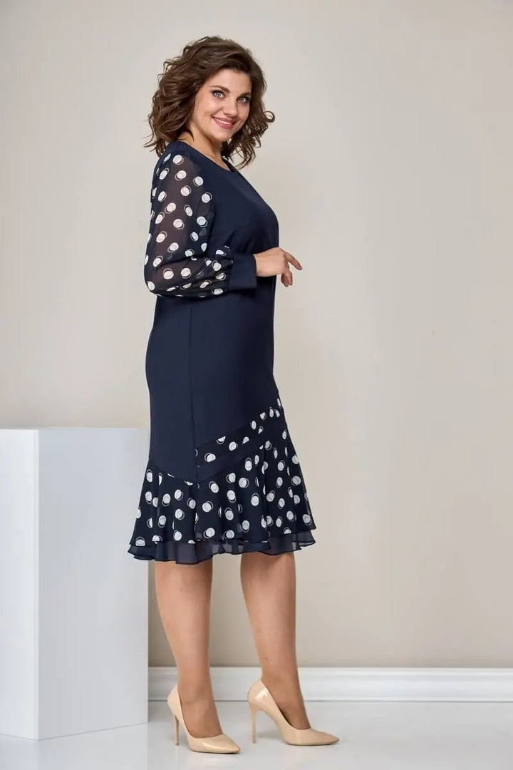Side view of woman in navy dress with polka dot sleeves, asymmetric ruffled hem, beige heels, in a neutral room.