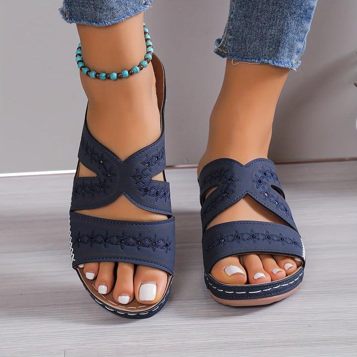 Front view of navy orthopedic sandals with decorative stitching, worn with frayed jeans, on a light wood floor.