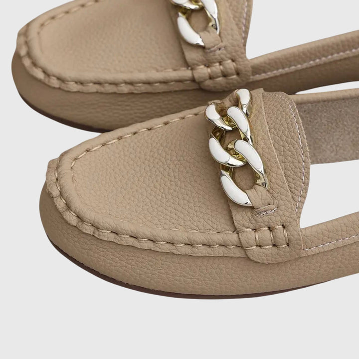 Close-up of beige orthopedic loafers with white and gold chain detail, on a gray background.