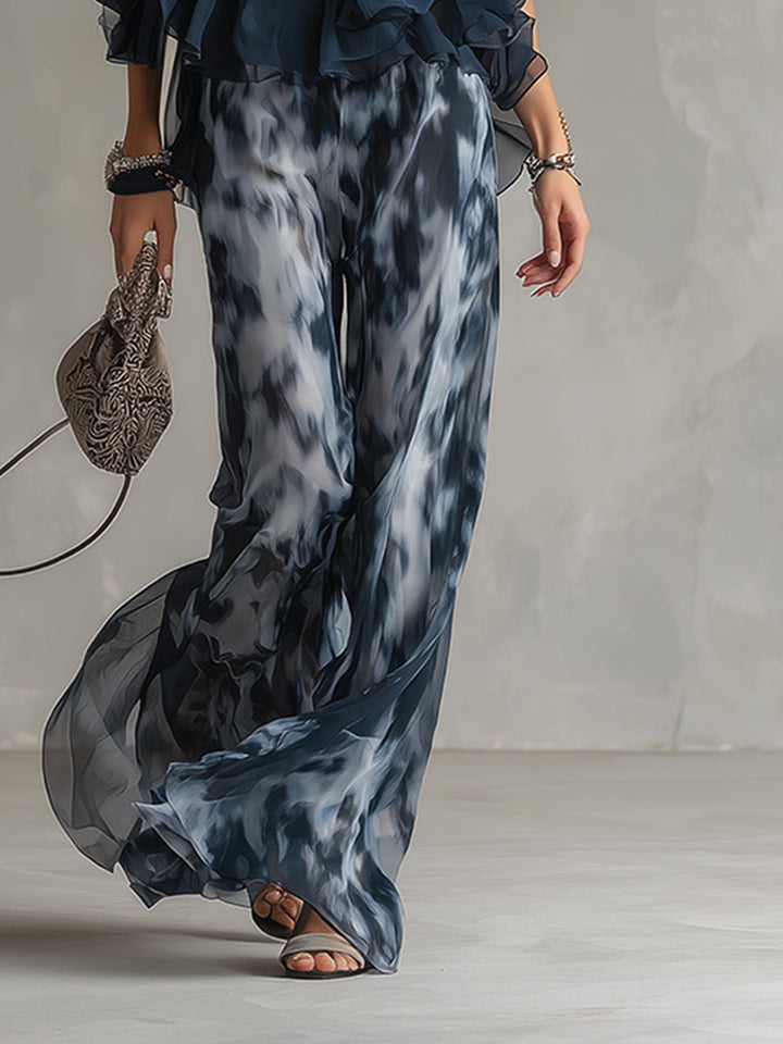 Close-up of wide-leg patterned pants and sandals, holding a delicate handbag. Studio setting.