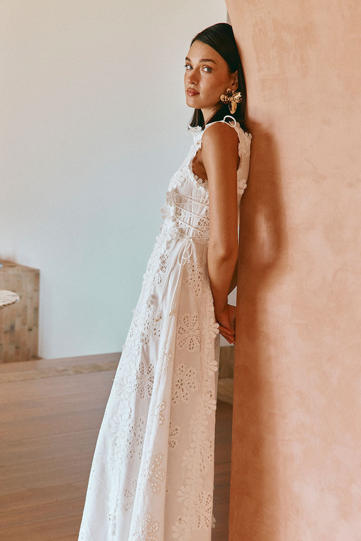 Woman in floral embroidered maxi dress leans against peach wall in softly lit room.