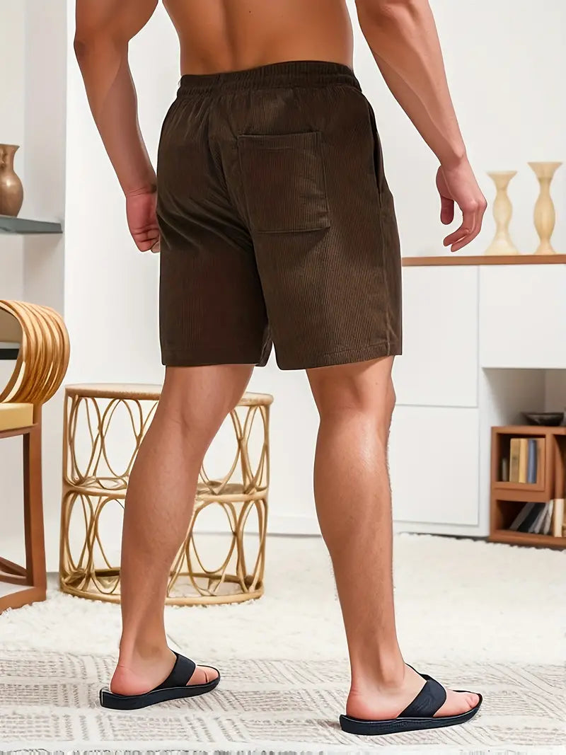 Back view of model in brown swimming trunks, standing in a cozy room with rug and furniture.