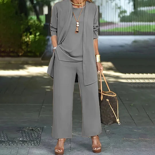 Gray three-piece set with relaxed top, long jacket, and wide-leg pants. Outdoor setting, woman carrying a handbag.