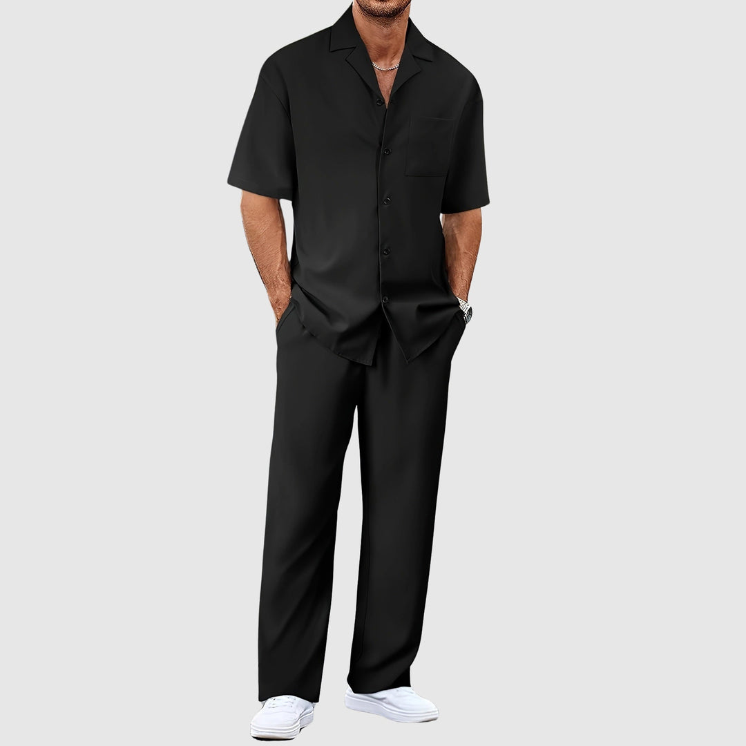 Man in black relaxed summer set with short-sleeve shirt and pants, hands in pockets, against gray studio background.