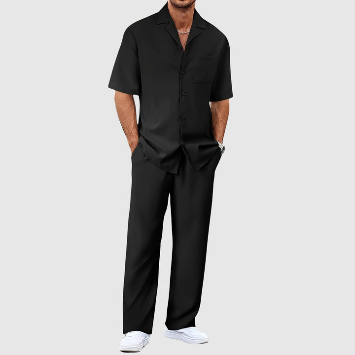 Man in black relaxed summer set with short-sleeve shirt and pants, hands in pockets, against gray studio background.