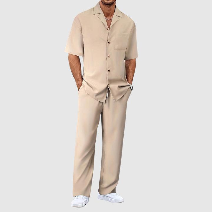Man in beige relaxed summer set with short-sleeve shirt and pants, hands in pockets, against gray studio background.