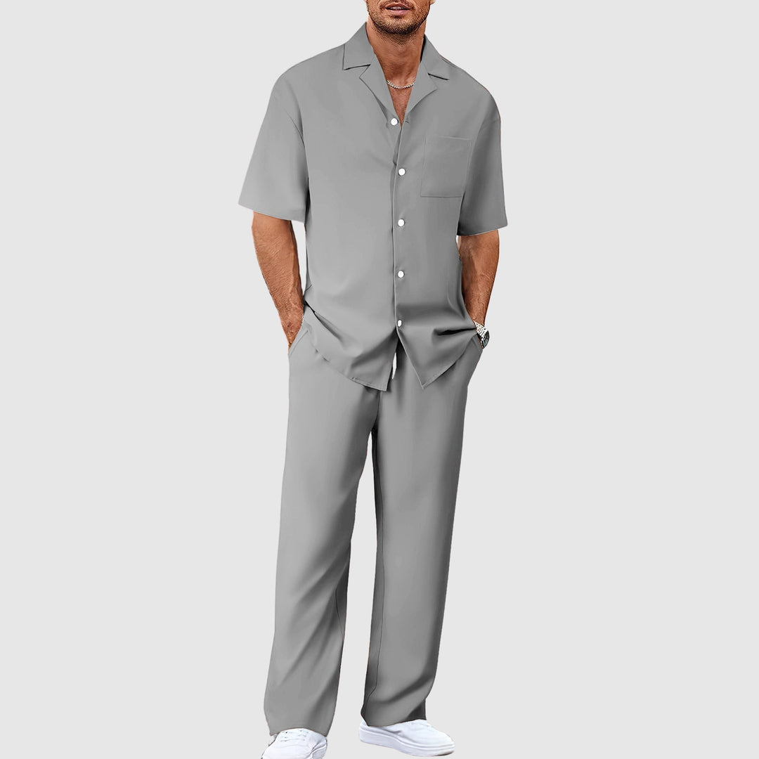 Man in gray relaxed summer set with short-sleeve shirt and pants, hands in pockets, against gray studio background.