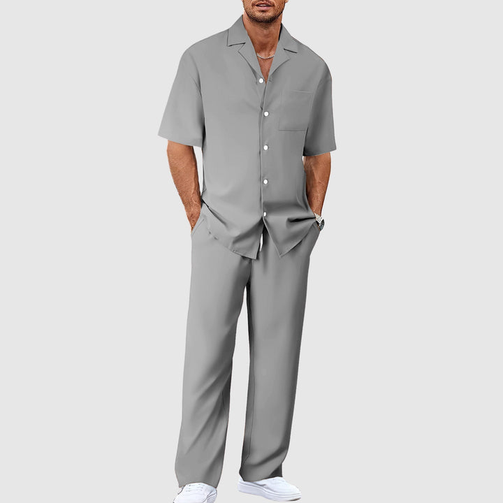 Man in gray relaxed summer set with short-sleeve shirt and pants, hands in pockets, against gray studio background.