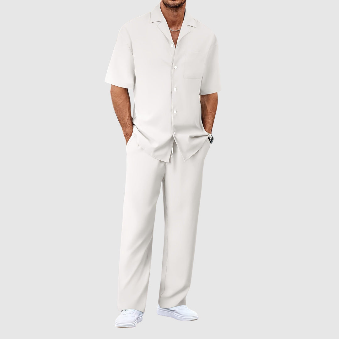 Man in white relaxed summer set with short-sleeve shirt and pants, hands in pockets, against gray studio background.