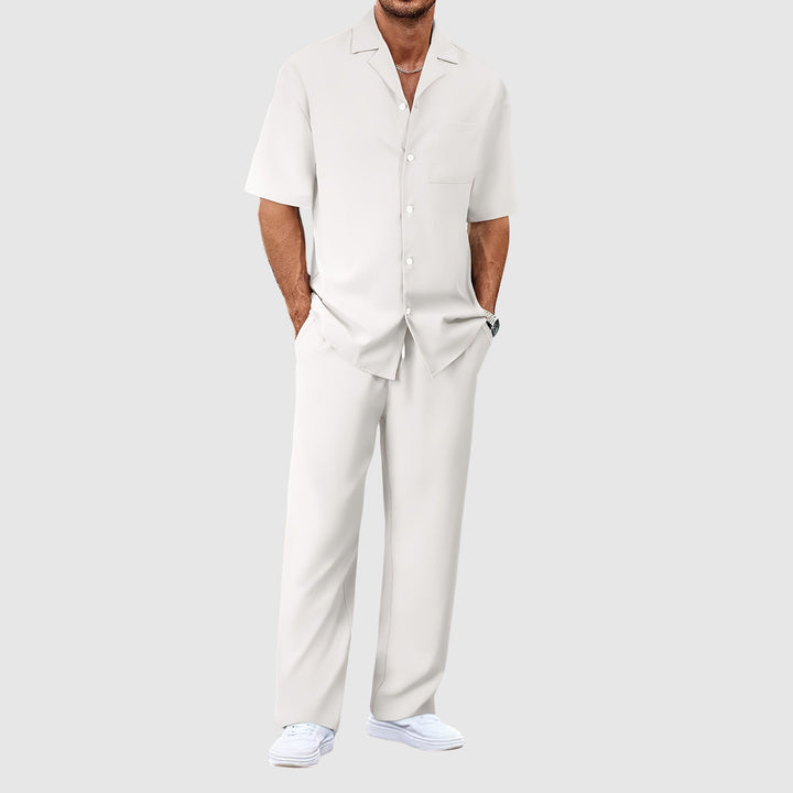 Man in white relaxed summer set with short-sleeve shirt and pants, hands in pockets, against gray studio background.