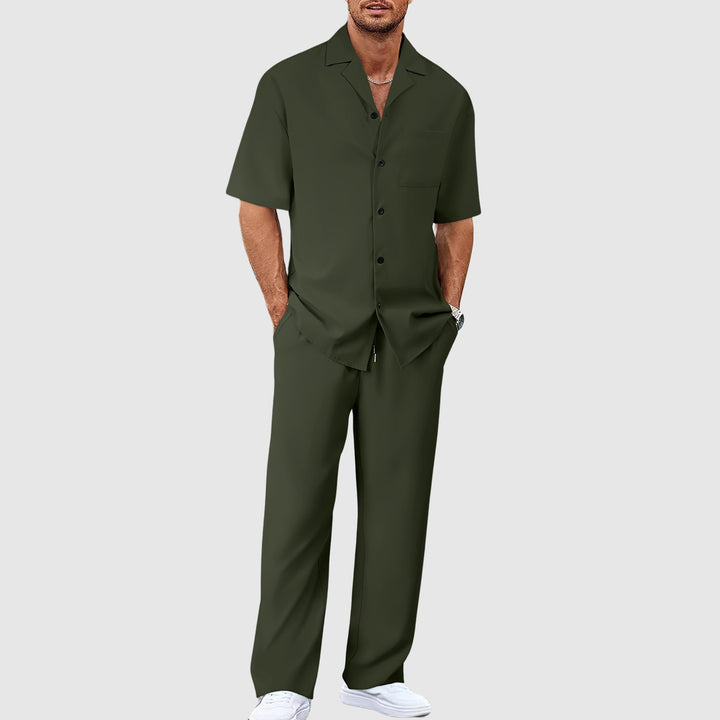 Man in dark green relaxed summer set with short-sleeve shirt and pants, hands in pockets, against gray studio background.