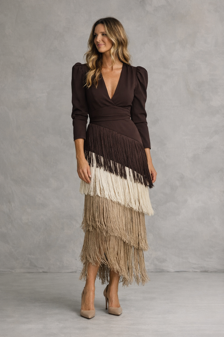 A woman in a brown and beige layered fringe dress, standing against a gray textured studio background.