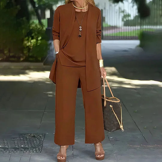 Brown three-piece ensemble with open jacket, top, and wide-leg pants. Setting is an outdoor path, woman holding handbag.