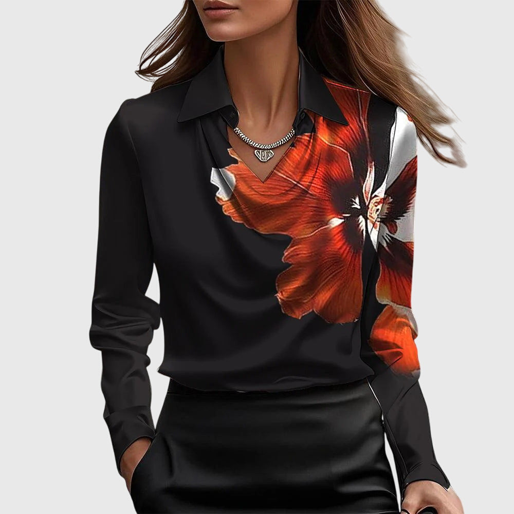 Black blouse with large red and white floral design, draped neckline, long sleeves, paired with a black skirt on a plain backdrop.