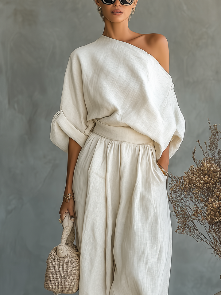 Upper body view of a woman in a one-shoulder white outfit holding a beige woven bag, next to dried flowers indoors.