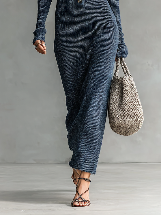 Lower portion of a model wearing a dark blue maxi dress holding a woven bag against a neutral background.