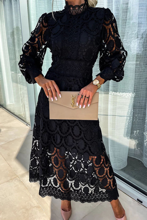 Black lace dress with intricate patterns, long sleeves, and high neckline, accessorized with a beige clutch. Studio setting with glass window backdrop.