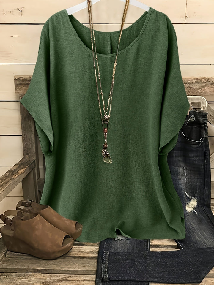 Green loose summer top on hanger with necklace, sandals, and jeans on rustic bench.