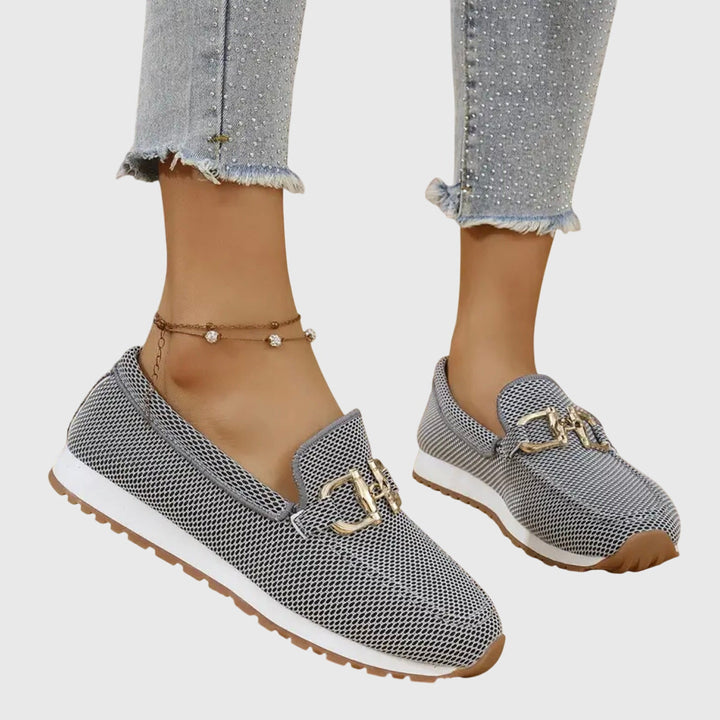 Gray orthopedic loafers with gold accents worn with frayed jeans and anklet on a light background.