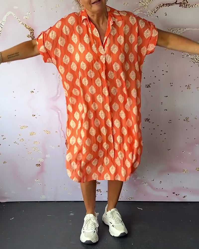 Model wearing an orange loose lapel collar dress, extending arms, styled with sneakers in a studio with abstract art wall.