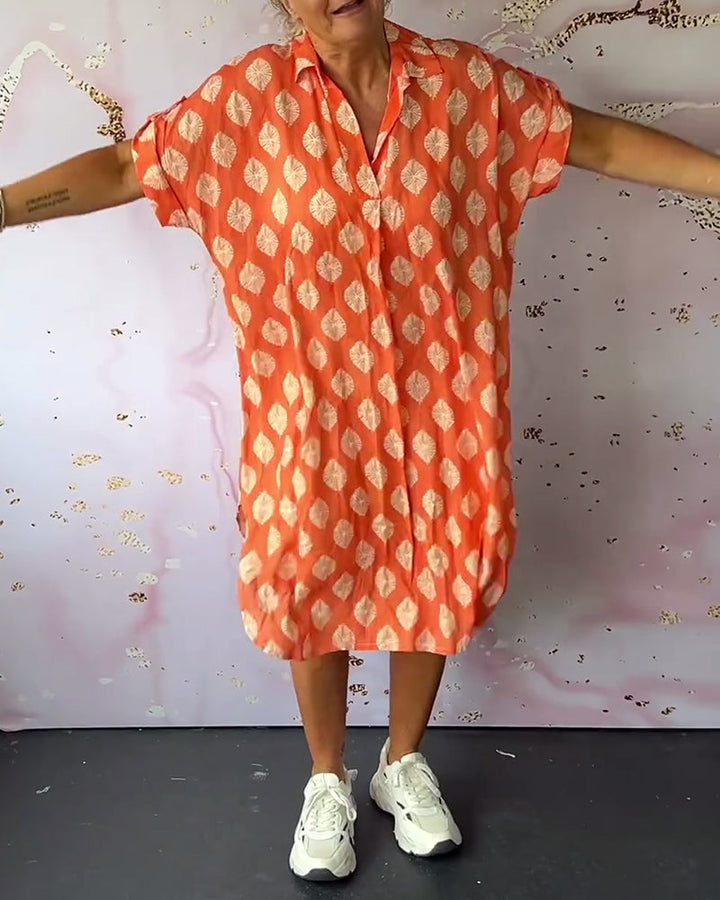 Model wearing an orange loose lapel collar dress, extending arms, styled with sneakers in a studio with abstract art wall.