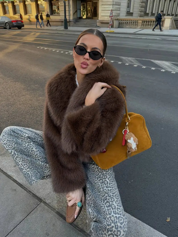 Person in a cozy brown fur jacket with leopard print pants, holding a yellow bag on a city street with historic buildings.