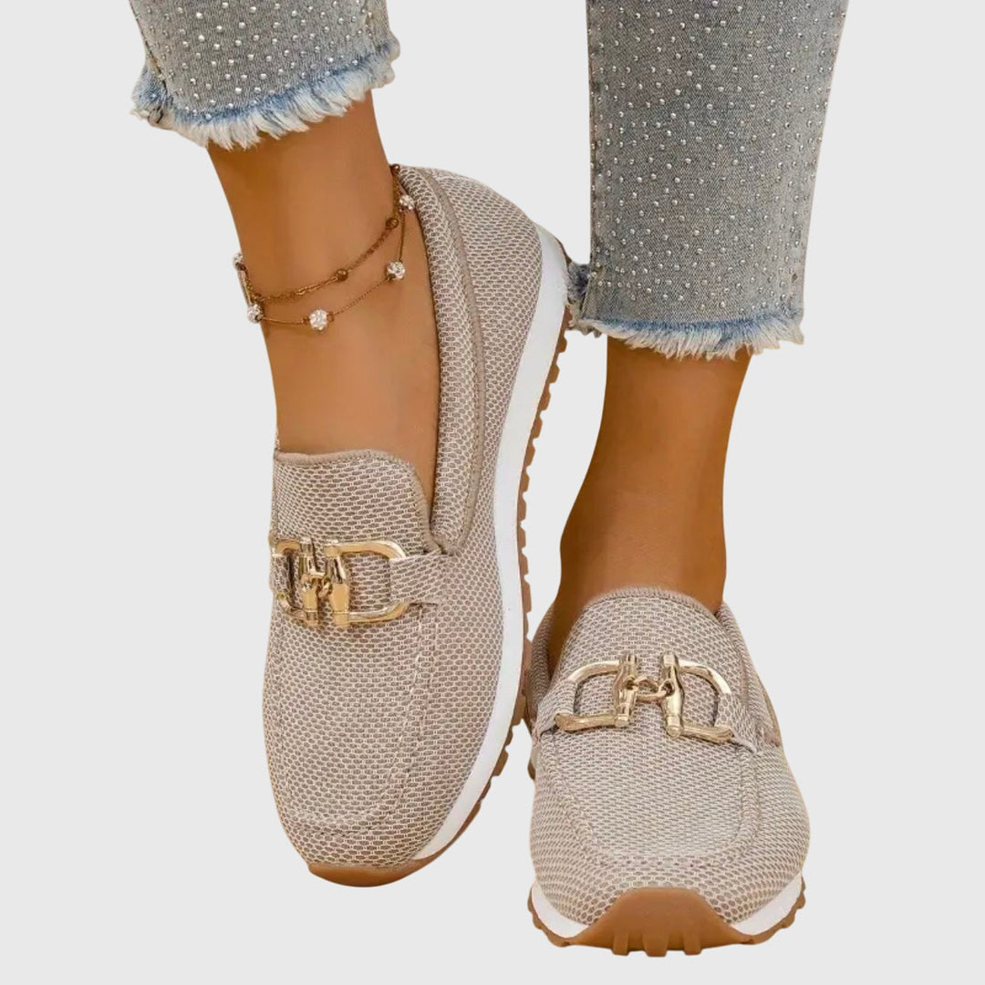 Pair of beige orthopedic loafers with gold embellishments worn with frayed jeans and anklet on light background.