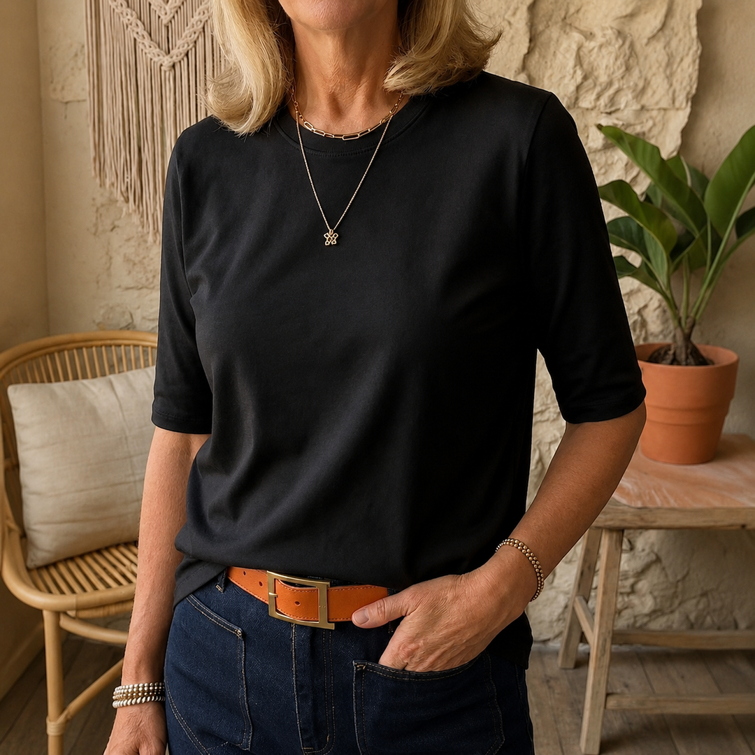 Black three-quarter sleeve tee with gold necklace, worn with denim jeans, in a cozy indoor setting with wicker chair and plant.