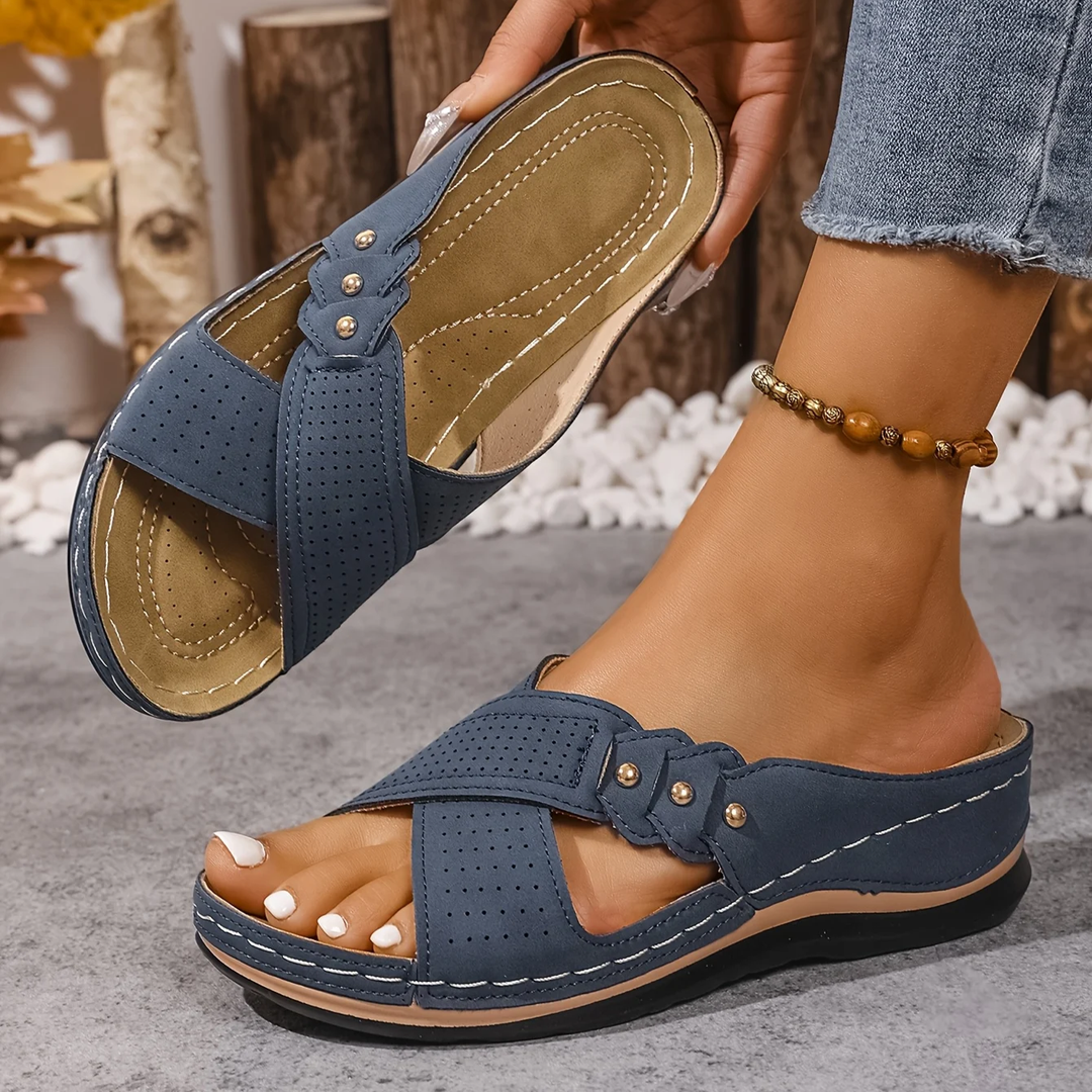 Blue orthopedic wedge sandals worn on feet, with wooden logs and white stones in the background.