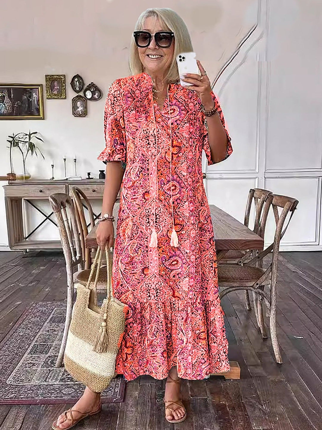Woman posing in lively boho dress with pink patterns, holding straw bag, in a stylish indoor setting with wooden elements.