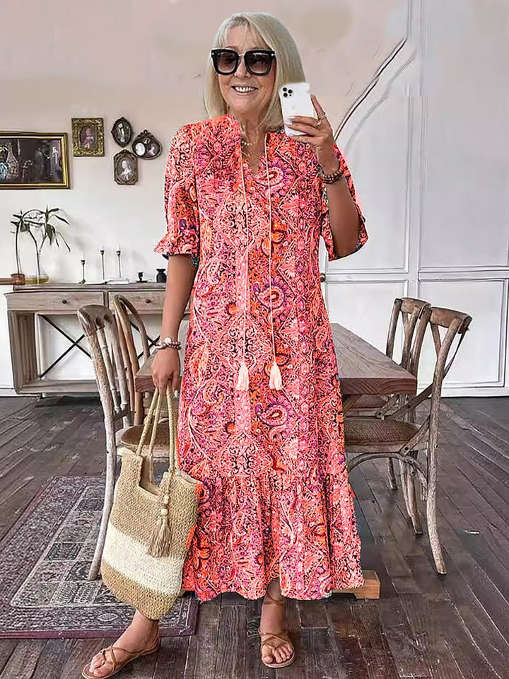 Woman posing in lively boho dress with pink patterns, holding straw bag, in a stylish indoor setting with wooden elements.